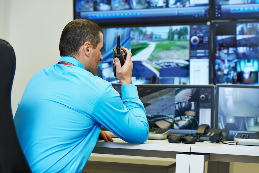 A man speaking into a walkie talkie while watching security camera feeds on multiple screens.