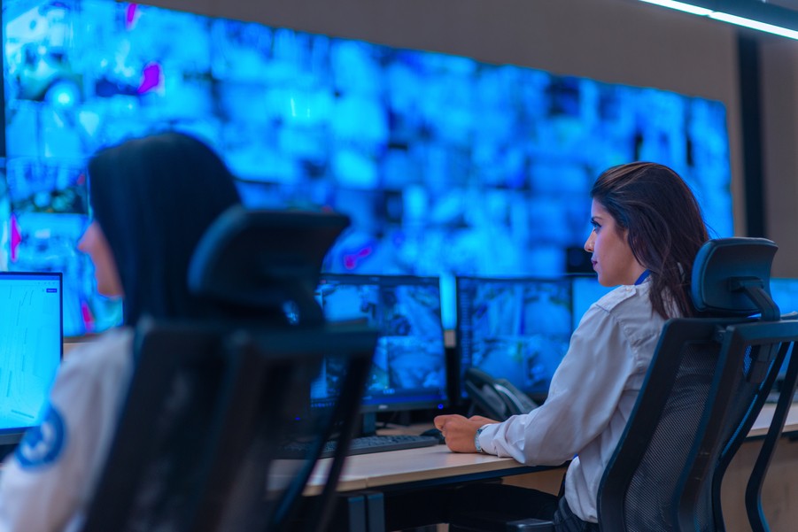 Two women monitoring a wall of screens, all depicting security footage.